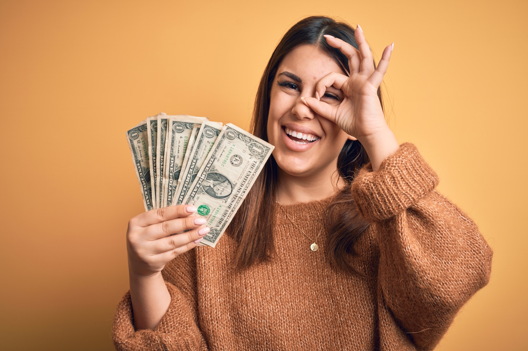 Young beautiful woman holding dollars standing over isolated orange background with happy face smiling doing ok sign with hand on eye looking through fingers