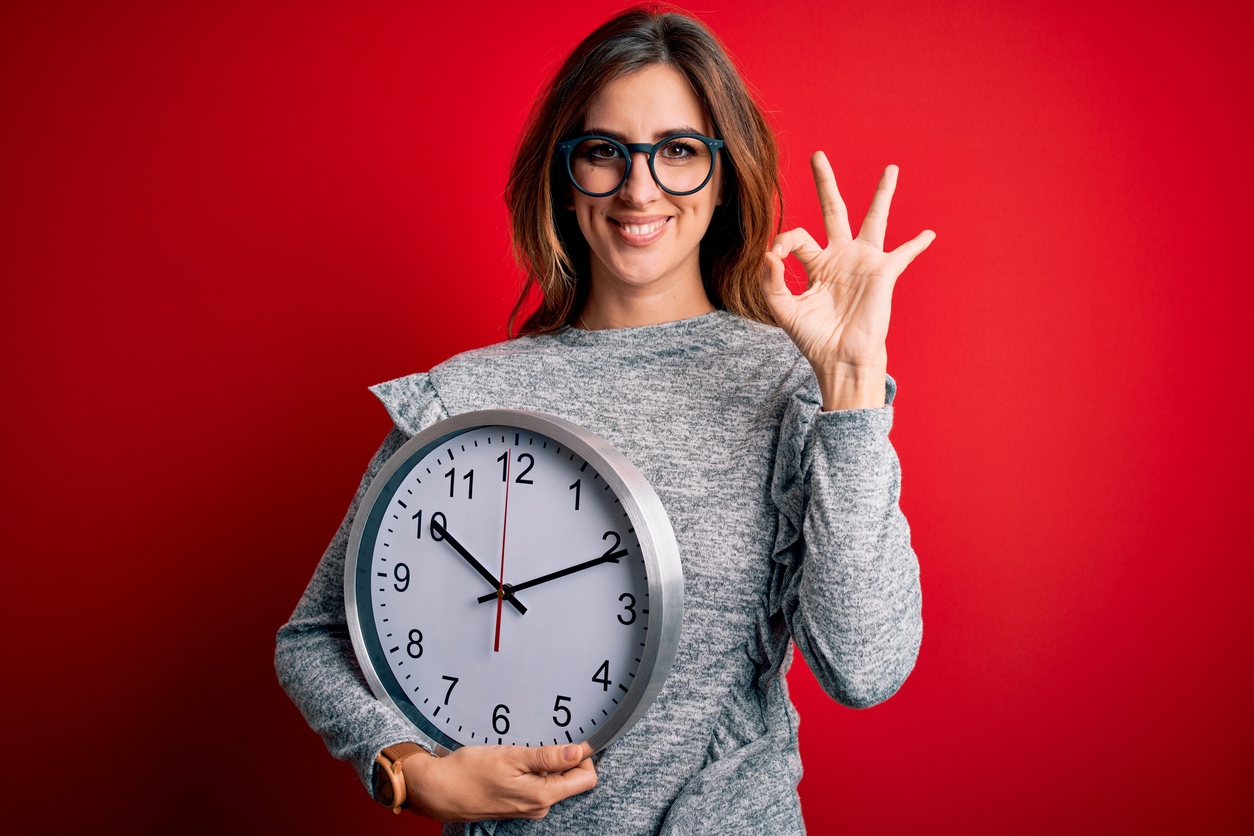 Young beautiful brunette woman doing countdown holding big clock over red background doing ok sign with fingers, excellent symbol