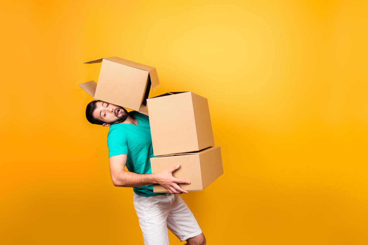 It’s moving day! Busy funny guy in casual clothes is carrying big boxes in his hands and trying to keep one using his head, isolated on yellow background